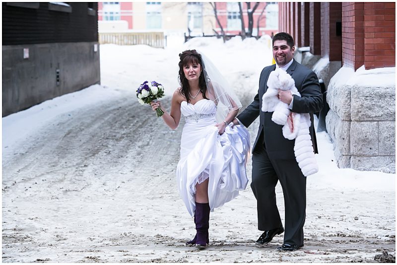 The groom holds up the wedding dress as they walk across a slushy street