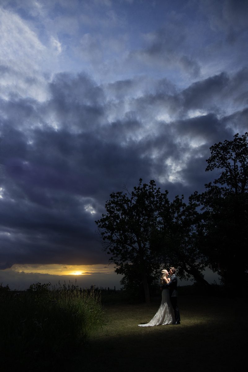 Bride and groom standing under a spotlight in a field with the sun setting in the background portrait