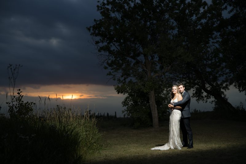 Bride and groom standing under a spotlight in a field with the sun setting in the background