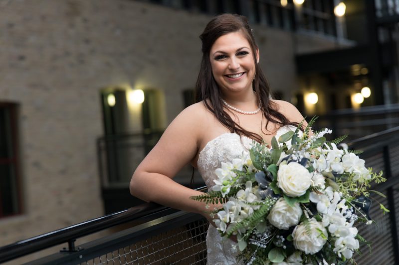 Jenn and big smiles while holding her custom bouquet leaning against a railing