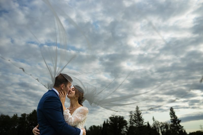 Arlita and Brendan kissing under amazing clouds with the veil floating across the sky