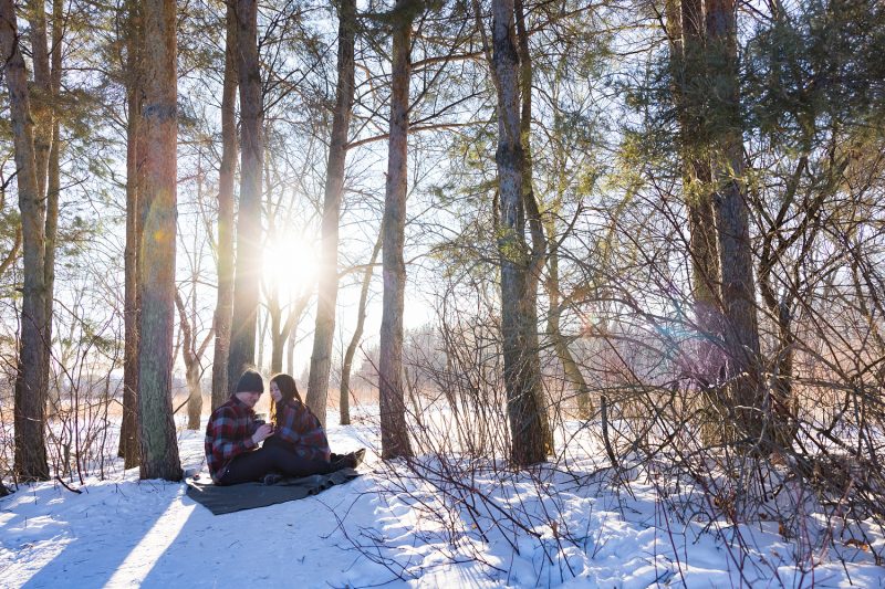 The couple sitting on a blanket on the snow drinking hot chocolate in the park in the middle of winter