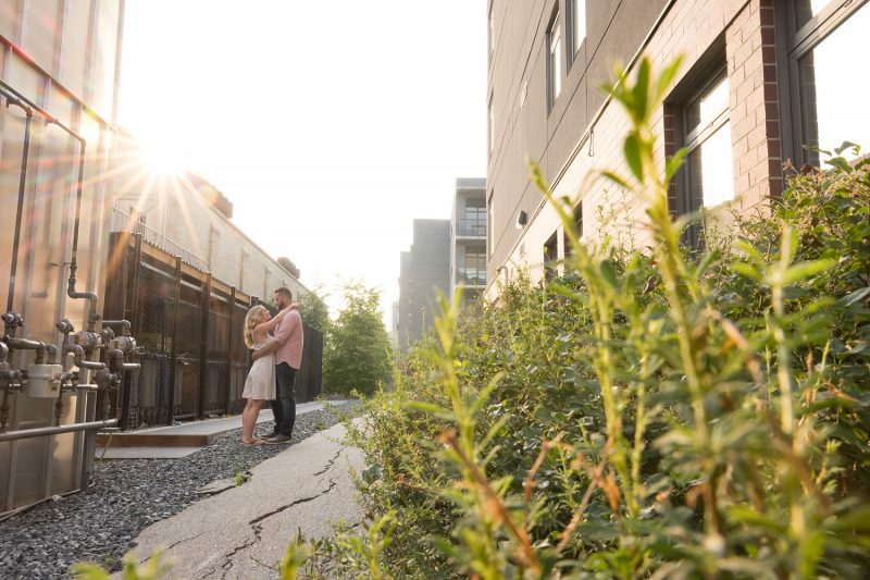 The couple holding each other in a back alley with the sun above the building