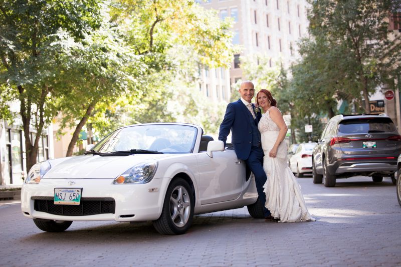 Trevor and Carrie embrace while they lean up against a white convertible car