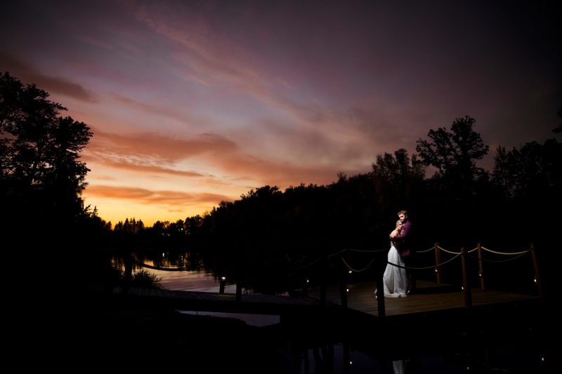 A dramatic, romantic photo of the couple being softly lit standing on a dock just before sunset with the sky lit up in oranges and the black tree line.