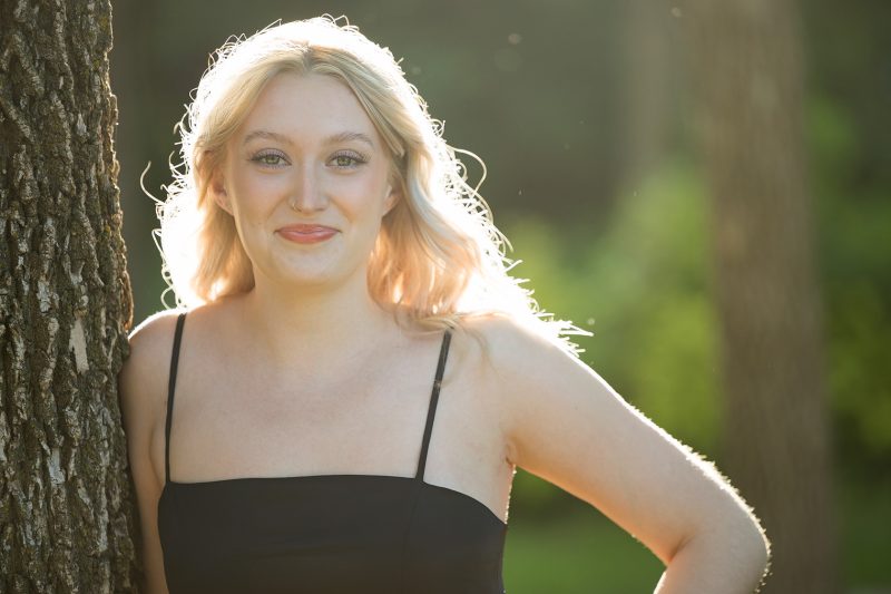 A young woman with long, blonde hair is smiling at the camera. She is wearing a black spaghetti-strap top and standing outdoors with a blurred green background.