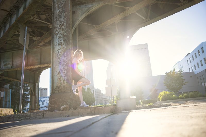 A person is leaning against a large, weathered metal column under an industrial structure with sunlight streaming through, casting a bright flare. The background includes an urban setting with buildings and greenery.