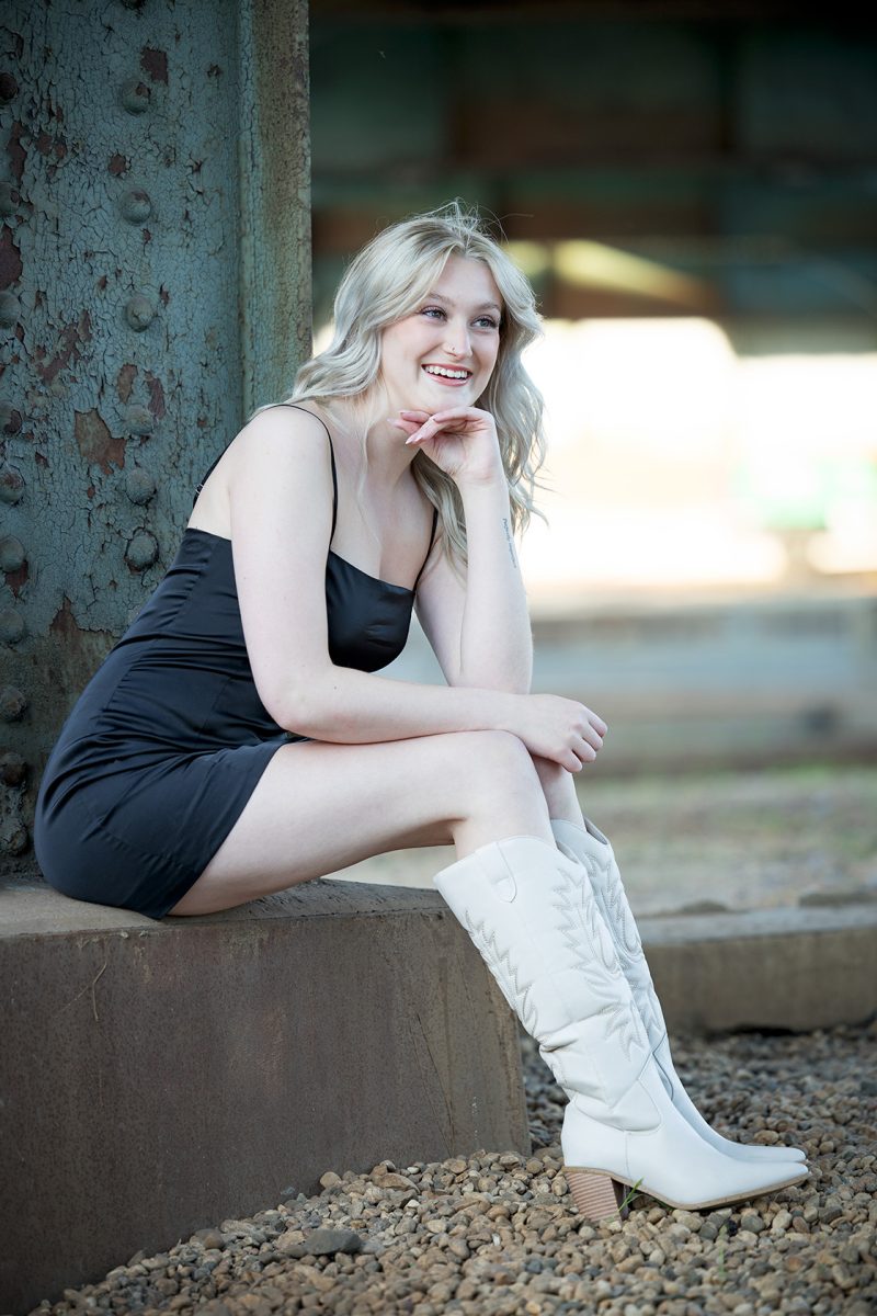 A young woman with long, blonde hair is sitting on a ledge and smiling. She is wearing a short black dress and white knee-high boots. The background appears to be an industrial or urban setting.