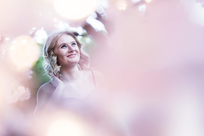 A photo of Brooklyn shot through some pink flowers with her wearing a big smile